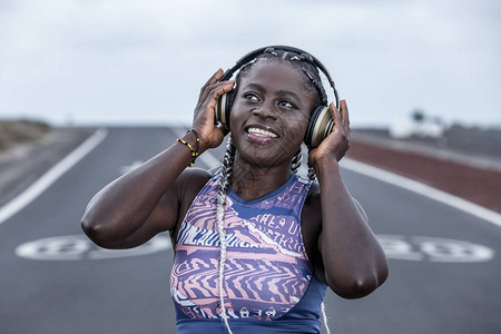 快乐的黑人女运动员在郊外户训练休息时调整耳机和微笑时站在图片