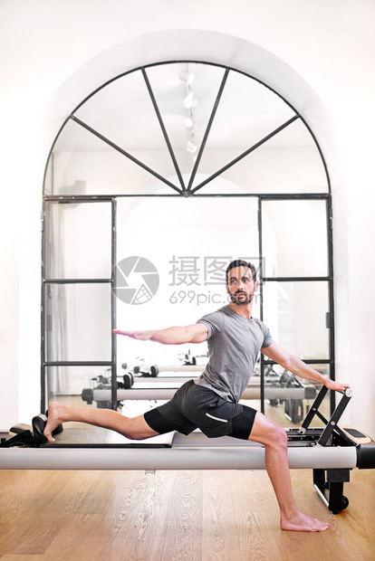 男子在健身房里使用改革者床做站立式普拉提弓箭步伸展运动图片