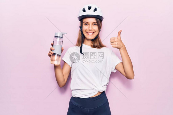 穿着自行车头盔的年轻漂亮女孩喝着一瓶水微笑快乐和积极拇指举起手表现优图片