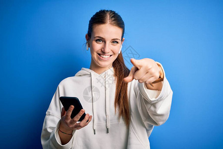 年轻的红发女运动员用耳机和智能手机指着你和用手指的相机听音乐图片