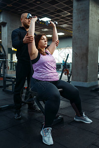 超重的女人与教练在体育俱乐部用哑铃锻炼图片