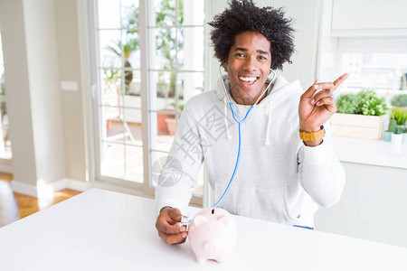 非裔美国人男子拿着听诊器检查小猪银行的金融健康图片