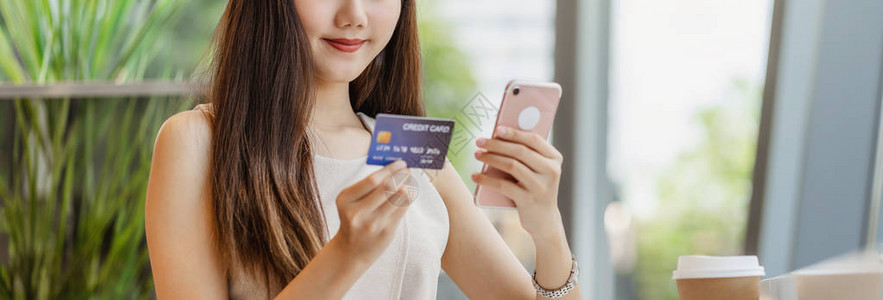 年轻的亚洲女使用带手机的信用卡在咖啡店或窗镜旁的合作空间进行网上购物技术货币钱包和在线支付概念背景图片