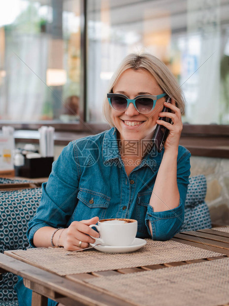 年轻漂亮的金发女郎在夏天的早晨戴着太阳镜在咖啡馆里用手机在一杯卡布奇诺咖啡上进行交流会议休闲早晨休图片