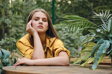 穿着黄衬衫的棕色头发年轻姑娘双手靠着头坐在城市公园绿叶图片