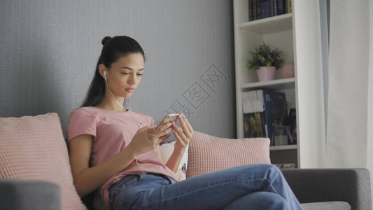 穿着粉红色枕头坐在灰色沙发上透过白耳机PHOTO收听智能手机音乐的年轻迷图片