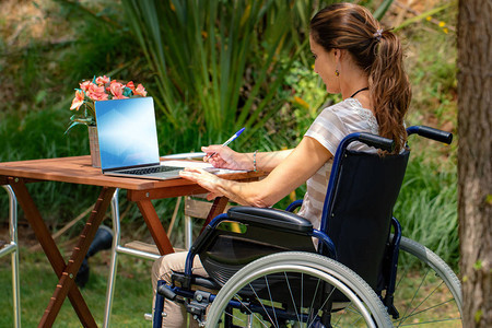在花园用笔记本电脑工作时坐在轮椅上的年轻瘫痪妇女的近距离肖像图片