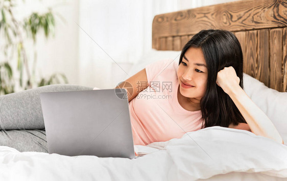 微笑的日本女孩在笔记本电脑上做远距离躺在家中卧床里工作图片