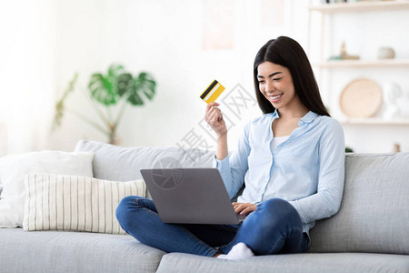 快乐的亚洲女在家使用笔记本电脑和信用卡在线购物坐在客厅的沙发上享受网上购图片