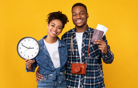 旅行时间微笑的黑人夫妇拿着机票和时钟准备度假站在黄色工作室背景旅行旅图片