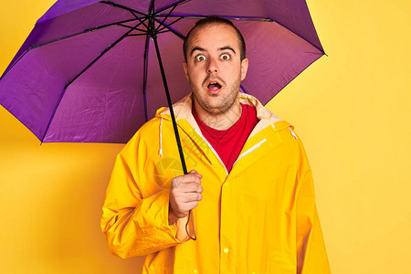 身穿雨衣手持紫色雨伞的年轻人站在孤立的黄色背景上图片