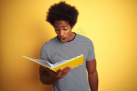 站在孤立的黄色背景上看书的美国黑人学生男子惊恐地惊讶地脸色惊恐图片
