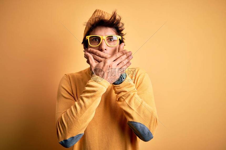身穿轻便T恤和眼镜的英俊男青年在孤立的黄色背景上被用双手遮住嘴顶着口图片