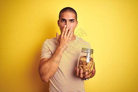 英俊的年轻帅哥拿着一罐饼干在黄色孤立的背景面罩上握着一只手掌图片