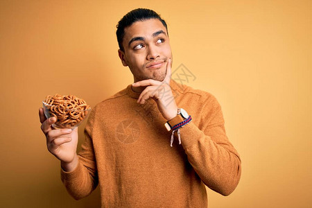 年轻巴西男子拿着碗和德国咸烤面包脆饼在黄色背景的背面严肃地思考问题图片