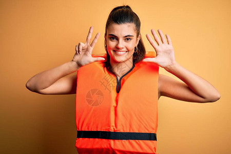 年轻美丽的黑发女人穿着橙色安全救生衣在黄色背景上显示并用八号手指向上图片