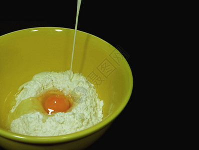 将牛奶倒入黑色背景的黄色陶瓷搅拌碗中的生面粉和蛋黄中图片