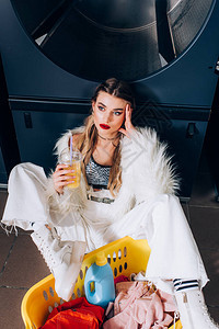穿着人造皮草夹克的年轻女子坐在洗衣店里的篮子附近图片