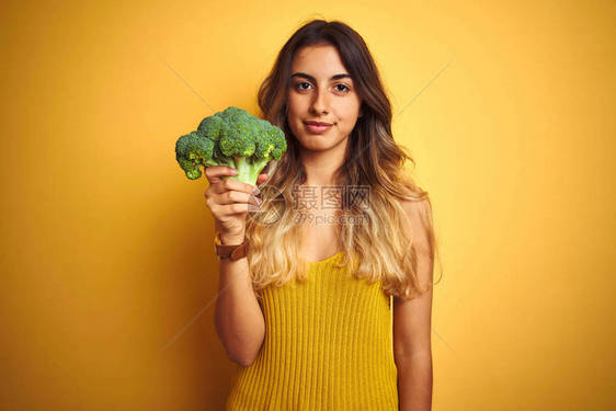 年轻美女在黄色孤立的背景上吃花椰菜并且自信地表现在聪明的面孔上图片