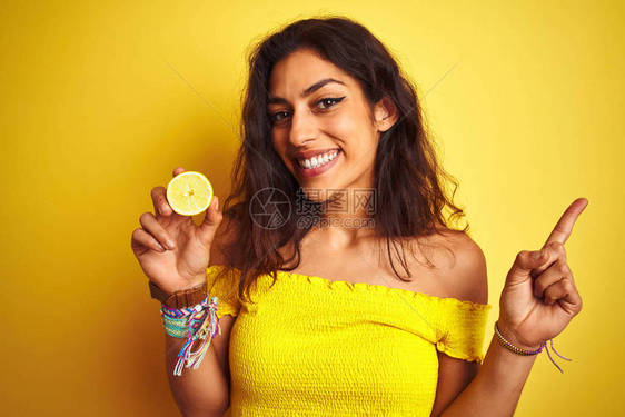 年轻美女拿着中间柠檬在孤立的黄色背景上站立快乐地用手和指对图片