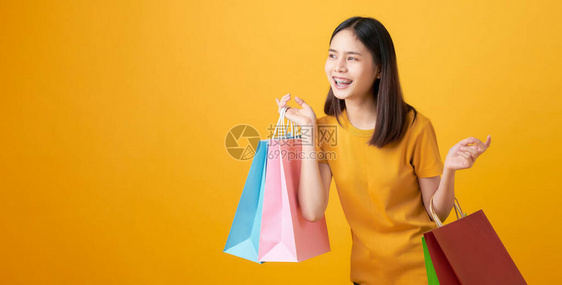 年轻笑容的亚洲女人拿着多色购物袋看着浅黄色的背景图片