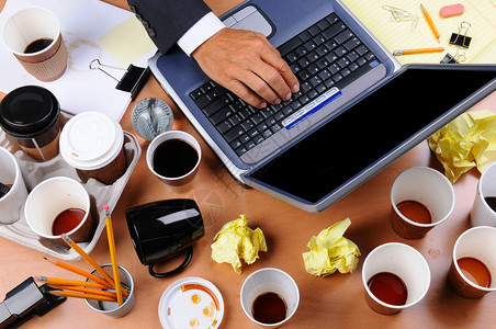 一个非常杂乱的商人办公桌的特写视图用人的手放在笔记本电脑键盘和散落的咖啡杯和办公用品上的俯视图片