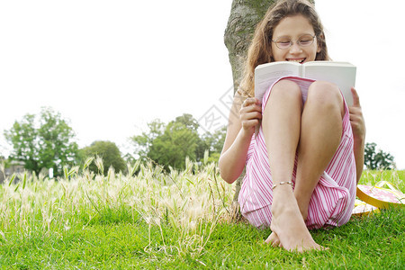 年轻女孩坐在公园的树下笑着看书的时候背景图片