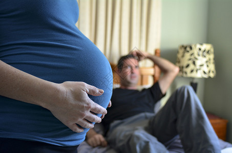 孕妇抱着她的腹部和困惑的男人躺在背景的双人床上图片