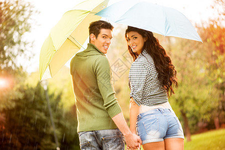看着一对爱着雨伞的年轻夫妇在雨中走过公园时举起手来图片