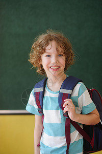漂亮的小学生站在黑板旁边的班级里在它的肩膀后面是一个书包学校生看着镜头微笑这个男孩有一头金色的卷发图片