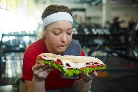 可爱的超重女在健身房锻炼时舔嘴唇想吃巨大的增肥三明治图片