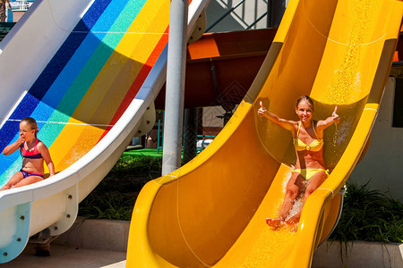 水上乐园蓝色水滑梯上的儿童游泳池滑梯夏季孩子们骑户外度假两件式泳装的快乐美丽姐妹女孩在水上公园举起图片
