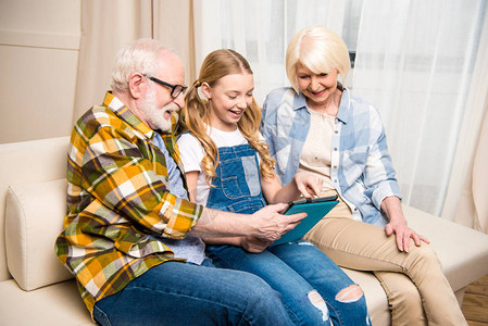 快乐的祖父母和微笑的孙女坐在沙发上使用图片