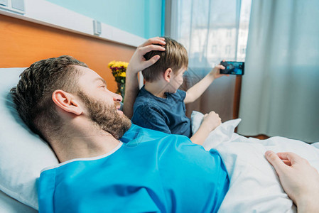 年轻父亲和儿子在病房用智能手机自拍图片