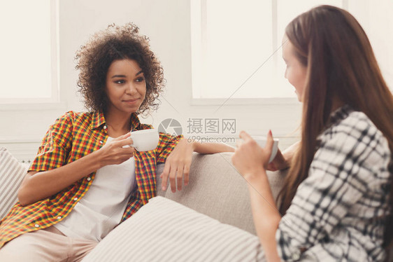 两个快乐的年轻女朋友拿着咖啡杯在家里的客厅里聊天图片