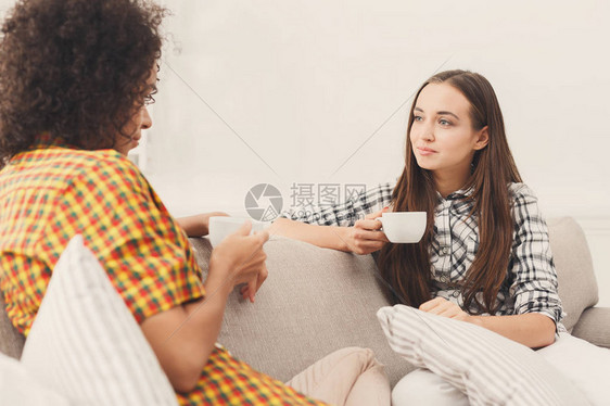 两个快乐的年轻女朋友拿着咖啡杯在家里的客厅里聊天图片