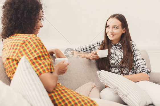 两个快乐的年轻女朋友拿着咖啡杯在家里的客厅里聊天图片