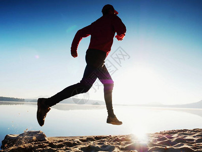戴着镜红色棒球帽和红黑色运动服的高个子男正在海滩上跑步和锻炼在山湖海滩上图片