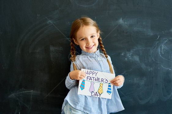 微笑的小女孩拿着手工制作的父亲节贺卡在学校的黑板上摆姿势看图片