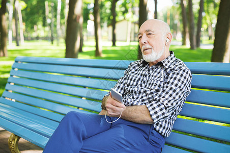 放松的老人在户外听音乐戴耳机的老人坐在公园的长椅上欣赏智能手机的音图片