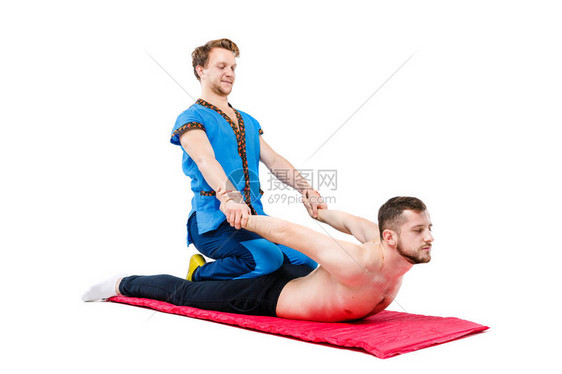 一位身着蓝色西装的年轻英俊男医生按摩师向一个躺在地毯上谋生的人摆出传统的takia按摩姿势白色孤立的背景图片