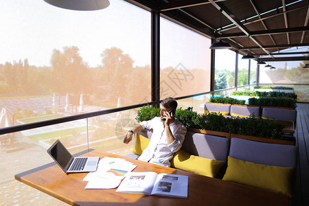成功的房地产经纪人在咖啡桌使用笔记本电脑并在智能手机上向客户输入信息英俊的高兴男坐在绿色室内植物附近人图片