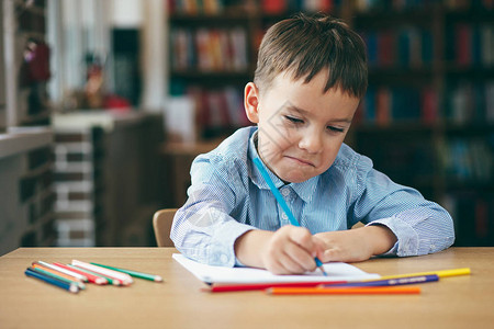 可爱的困惑微笑男孩做作业着色页写作和绘画孩子们画孩子们画图书馆里有书的学龄前儿童桌子上的彩色铅笔和纸背景图片