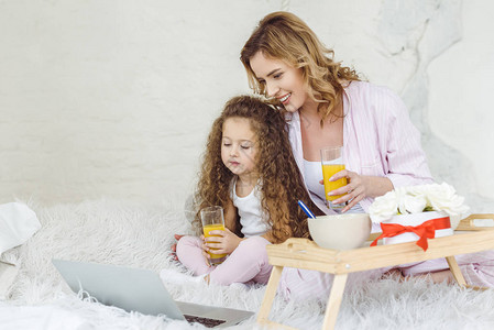 母亲和女儿用笔记本电脑在床上用橙汁杯子图片