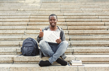微笑坐在楼梯上举起拇指用笔记本电脑工作准备大学校园考试的非洲美籍学生图片