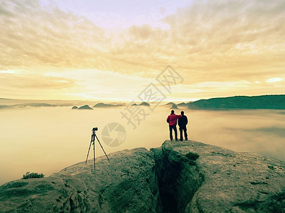 两位暖和的大衣摄影师带着三脚架在山下拍摄秋天的风景多云的山上多云的雨图片