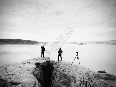 两位暖和的大衣摄影师带着三脚架在山下拍摄秋天的风景多云的山上多云的雨图片