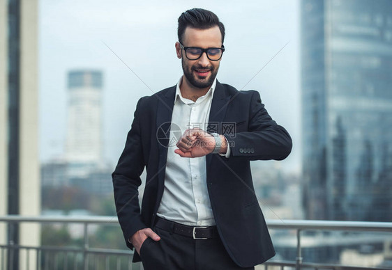 穿着西装和戴眼镜的英俊商人站在阳台上看着他的手表图片