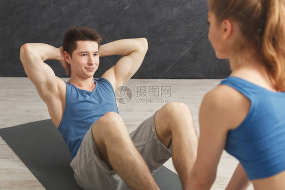 健身教练帮助在室内为年轻运动员做仰卧起坐年轻的运动型男人和女人在垫子上做仰卧起坐健康生活方式体图片