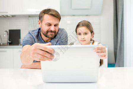 使用笔记本电脑和女儿坐在现代厨房的桌子上坐着时笑着的胡图片